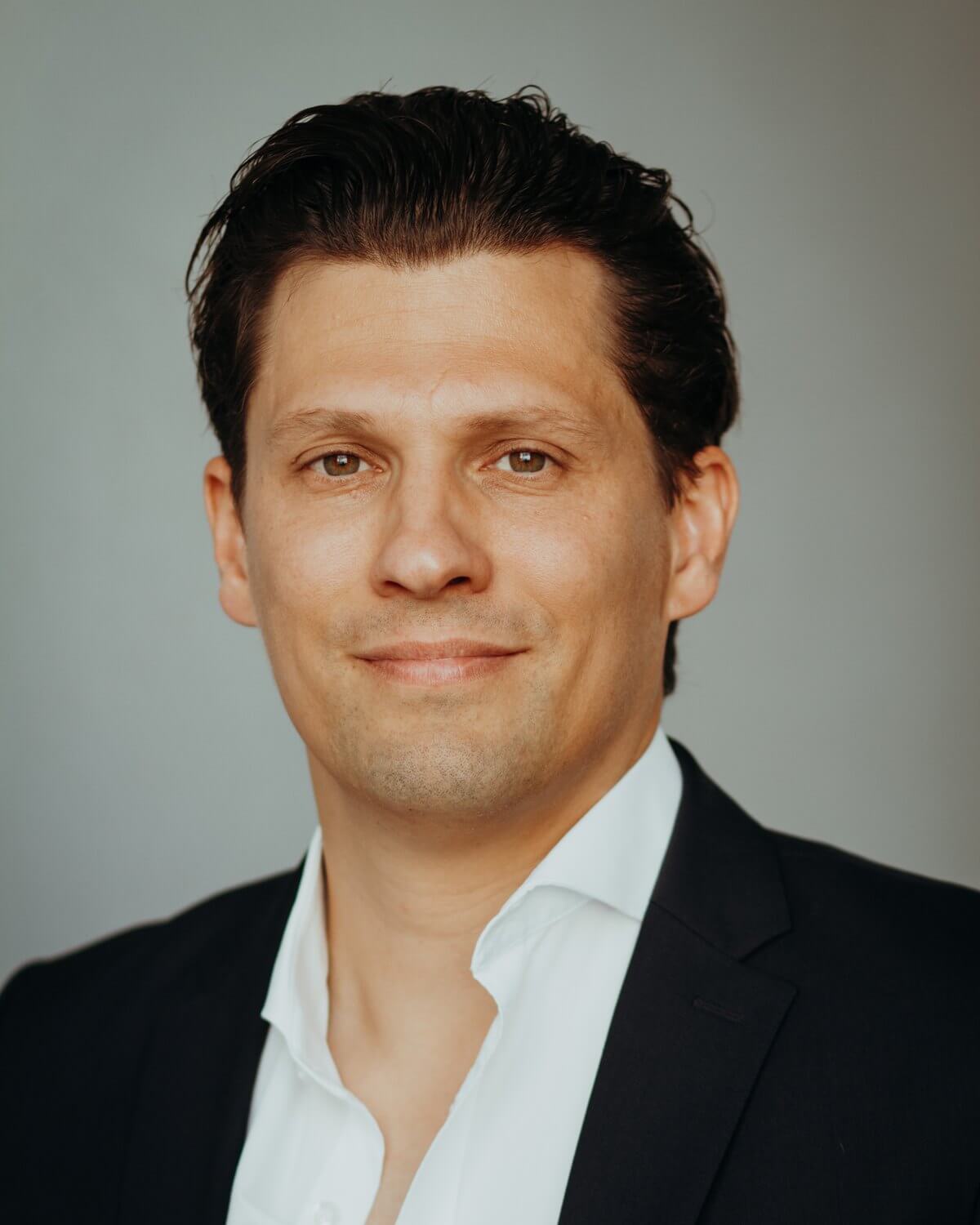 Porträt von Dr. Hubertus Porschen im dunklen Sakko mit offenem weißem Hemd vor hellgrauem Hintergrund