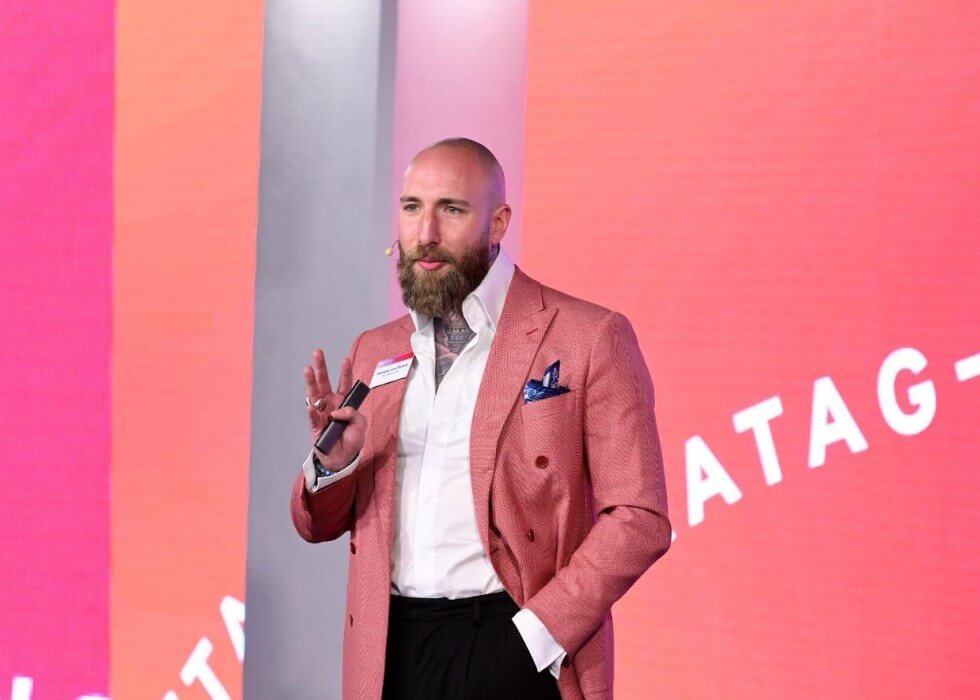 Dominic von Proeck mit Mikrofon auf einer Bühne vor pinkfarbenem Bühnenbild