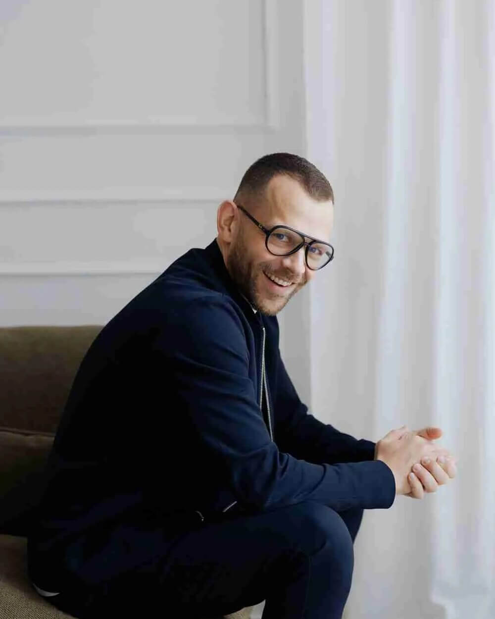 Christian Schlicht im dunkelblauen Blouson, seitlich auf einem Sofa sitzend vor einem hellen Vorhang